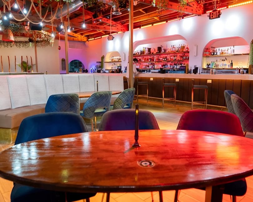 A vibrant bar interior with red lighting, a well-stocked bar, and empty tables with plush chairs.