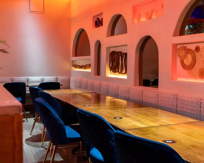 Restaurant interior with long wooden tables, blue velvet chairs, and arched wall niches with decorative items.