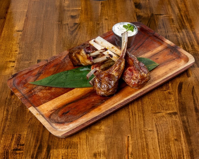 Grilled lamb chops with a side of white sauce and a green leaf on a wooden serving board.