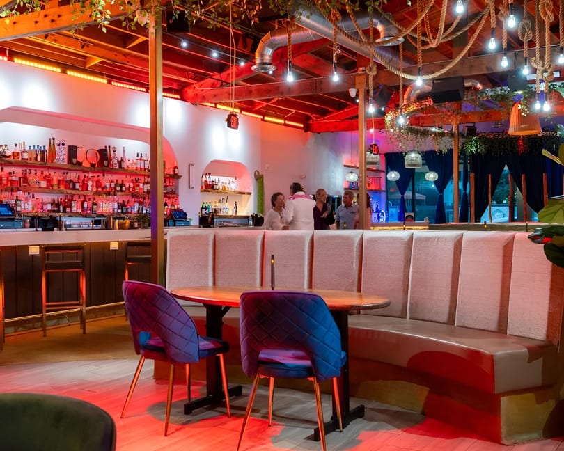 Restaurant interior with red lighting, a full bar, and guests conversing near booth seating.