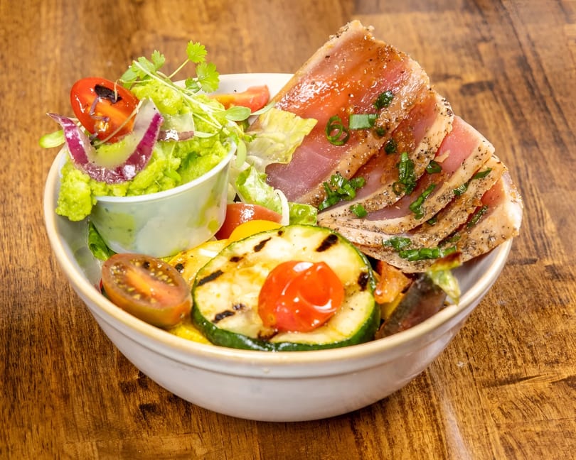 Seared tuna steak with grilled zucchini, cherry tomatoes, and guacamole in a white bowl.