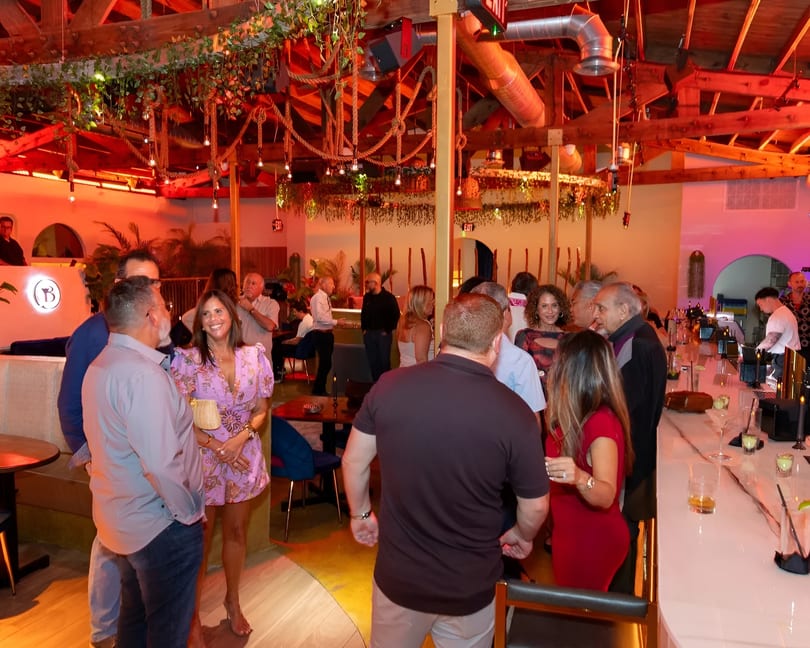 Patrons socialize in a vibrant restaurant with exposed wooden beams, hanging plants, and a lively bar area.
