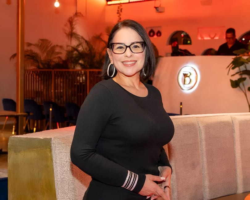Smiling woman in black top and glasses, with a restaurant interior and DJ booth in the background.