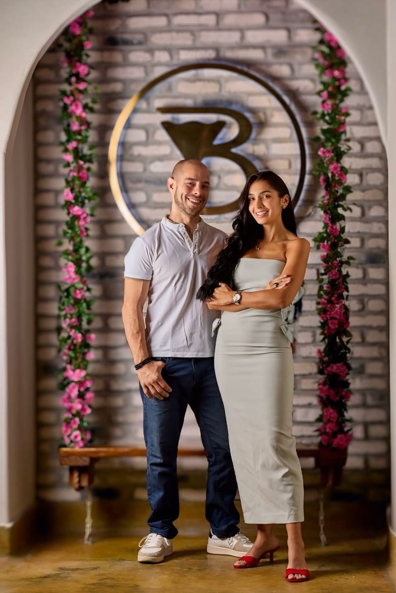Smiling couple standing in front of a brick wall with a golden logo and pink floral decor.