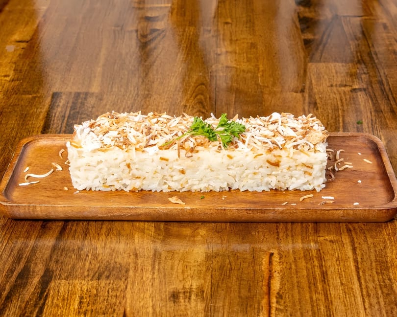 A rectangular serving of white rice topped with toasted coconut flakes and a sprig of parsley.