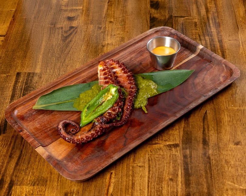 Grilled octopus with green sauce, a halved jalapeño, and a side of orange sauce on a wooden tray.