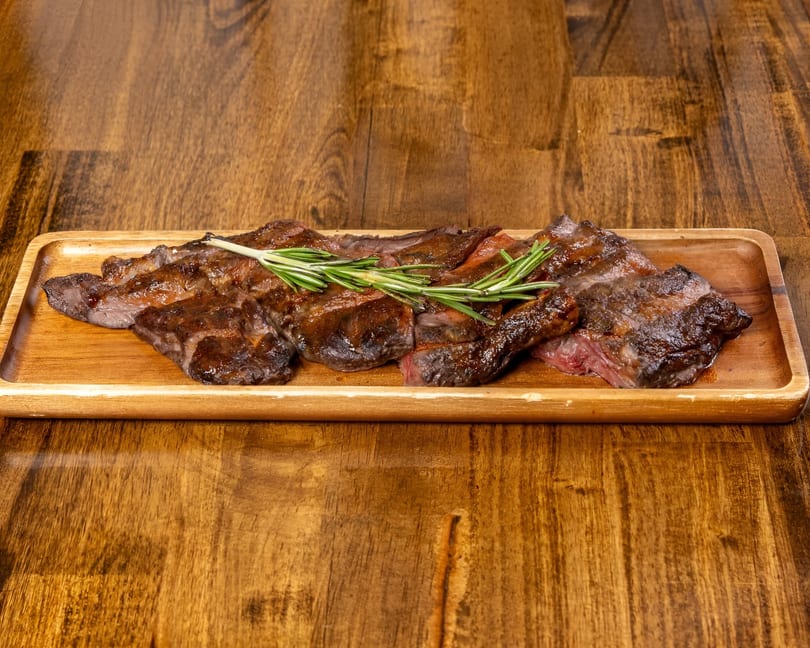Sliced steak with rosemary sprigs on a wooden serving tray, against a wooden table.