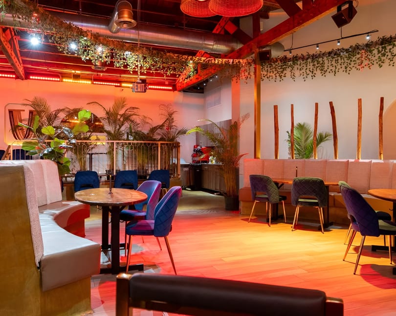 Restaurant interior with red lighting, green plants, and blue and green chairs around wooden tables.