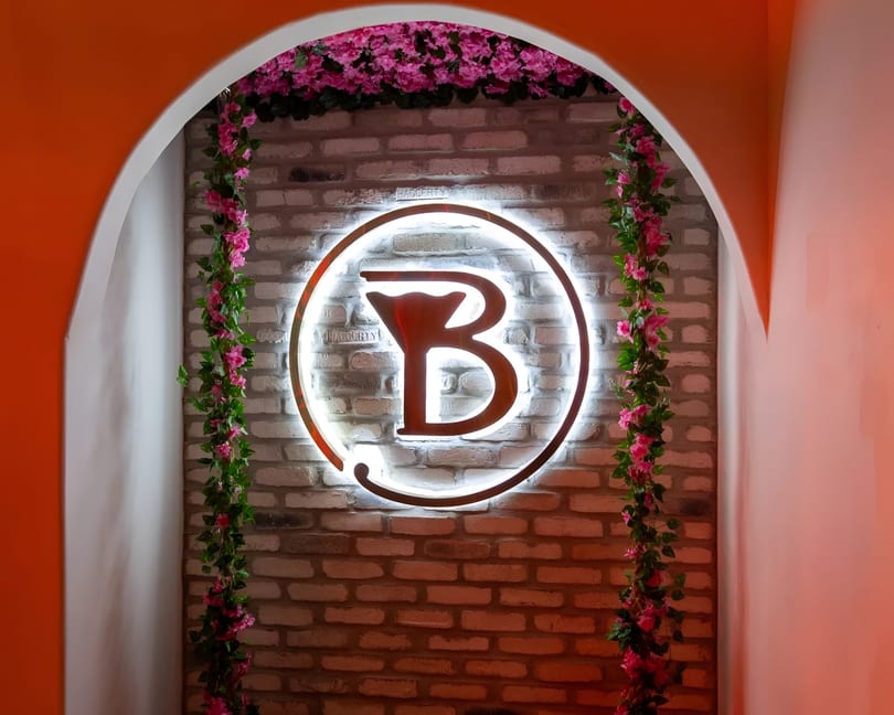A glowing 'B' logo with a martini glass icon, set against a brick wall with pink floral vines.