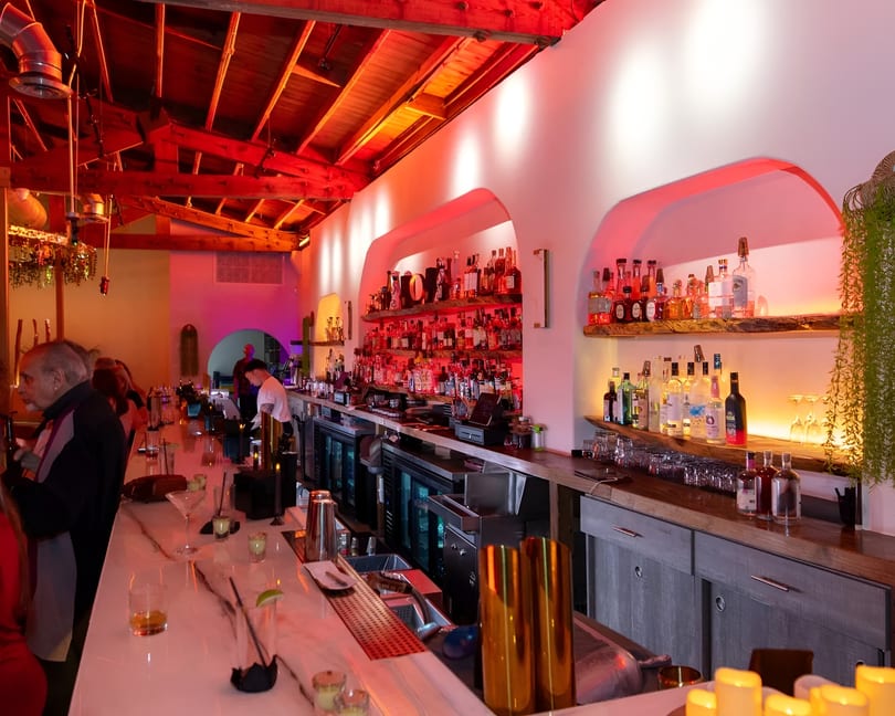 A vibrant bar with red lighting, a long white counter, and shelves filled with liquor bottles.