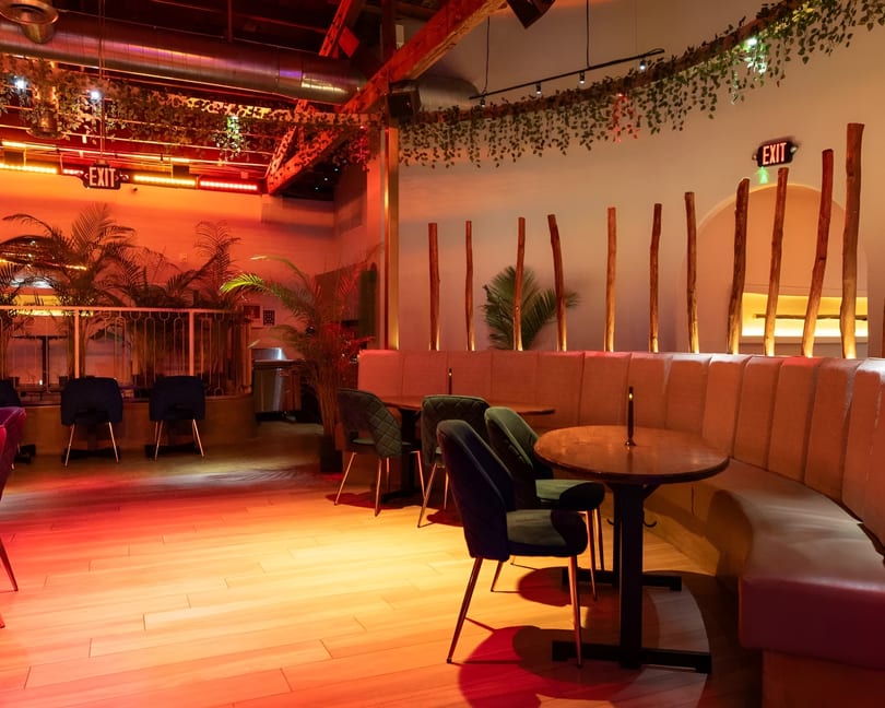 Restaurant interior with red lighting, green chairs, wooden tables, and tropical plants.